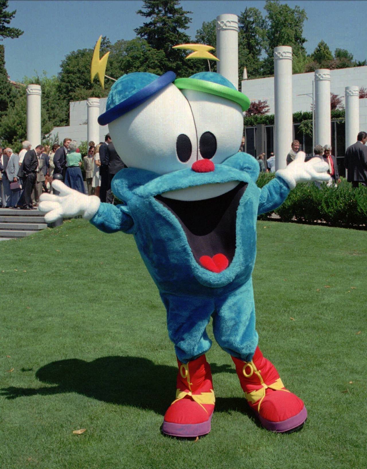Mascota Izzy de color azul, ojos grandes y zapatos anaranjados en un parque sonriendo